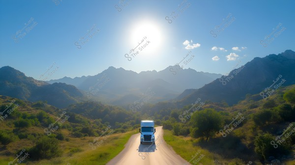 A large truck driving on a narrow road in a green valley surrounded by rocky hills and mountainous landscape. The sun is shining in the clear blue sky, illuminating the background and casting shadows over the hills. A few small white clouds are visible in the upper right corner of the image.