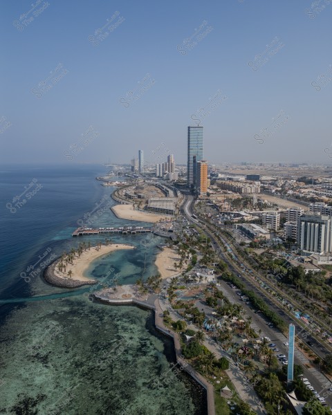 Aerial view of Jeddah Corniche in Saudi Arabia. The image shows the Red Sea on the left and the corniche stretching along the coast. Several modern buildings and skyscrapers are visible to the right, along with trees and green areas in the foreground. The water appears clear blue, surrounded by sandy beaches. The sky is blue and clear.