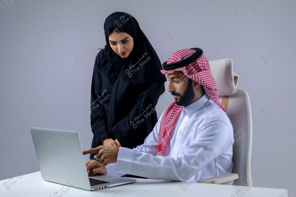 صورة لرجل وامرأة يرتديان ملابس تقليدية سعودية, حيث يجلس الرجل على كرسي ويستخدم جهاز كمبيوتر محمول. يرتدي الرجل ثوبًا أبيض وغترة حمراء وبيضاء وعقالًا أسود. تقف المرأة بجانبه وترتدي عباءة سوداء وحجابًا أسود, وتشاركه بالنظر إلى شاشة الكمبيوتر. يبدو أن كلاهما يشارك في عمل أو مناقشة مهنية.