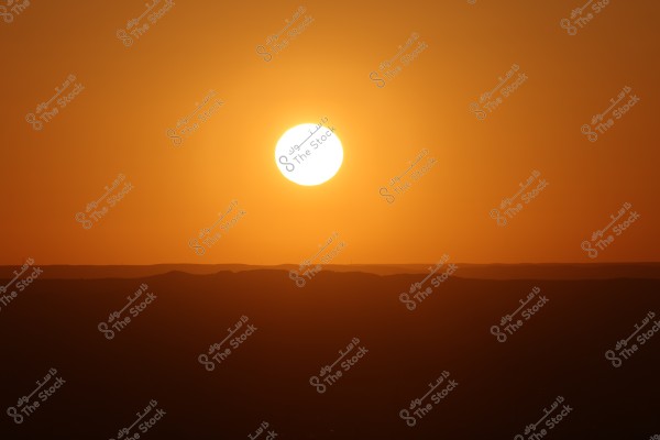 صورة لغروب الشمس فوق تلال, حيث تملأ الشمس الكبيرة مشهد السماء بلون برتقالي دافئ. تظهر الأفق بشكل غير واضح في الظلام, مما يضفي إحساسًا بالهدوء والجمال الطبيعي.