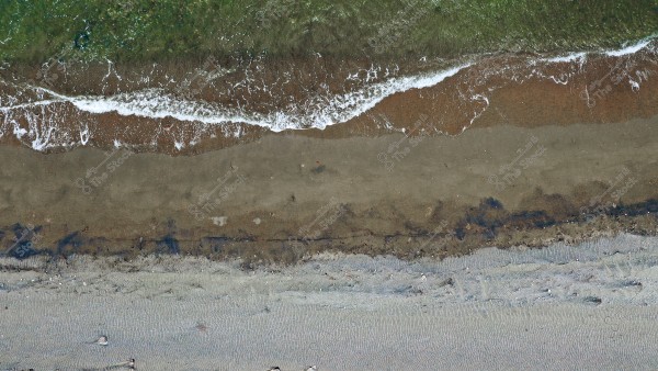 Aerial view of a sandy beach with ocean waves crashing onto the shore. The beach shows a contrast of colors between the green-blue water and the brown sand, with some scattered rocks visible on the shore.