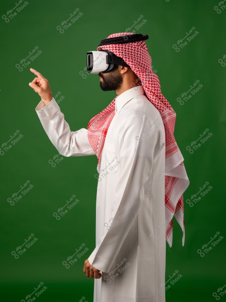 رجل يرتدي زيًا سعوديًا تقليديًا ونظارة واقع افتراضي يقف في وضع يشير إلى الأمام, على خلفية خضراء.