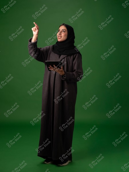 An image of a woman standing against a green background, wearing a black abaya and hijab. She is holding a tablet in her left hand and pointing upwards with her right hand, smiling.
