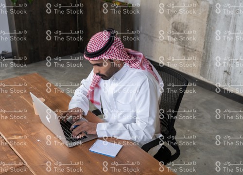 رجل سعودي يرتدي الزي السعودي التقليدي يجلس في مكتب و يستخدم الابتوب وبجانبه دفتر و كوب، موظف يعمل في المكتب