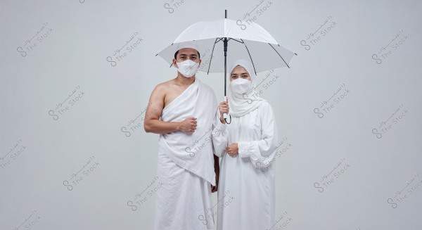 An image depicting a man and a woman in traditional white ihram clothing; the man is wearing two towels with his left shoulder covered, while the woman is wearing a white abaya and hijab. Both are wearing white medical masks, and the woman is holding a white umbrella. The background is light gray.