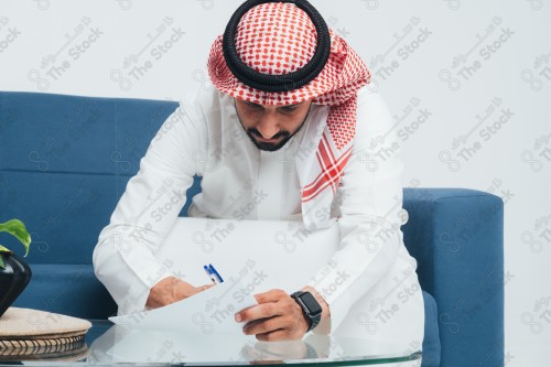 رجل سعودي يرتدي الزي السعودي التقليدي يجلس على أريكة ويراجع ورقة عمل