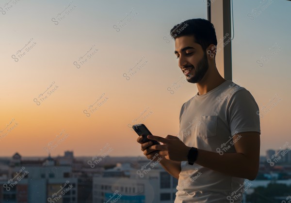 A photo of a man standing by a window browsing his mobile phone, smiling, wearing a simple t-shirt and a wristwatch. Behind him is a cityscape at sunset, with the sky filled with a warm orange hue.