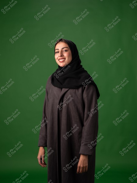 A portrait of a woman wearing a black hijab and a dark long coat, smiling at the camera. The background is green, highlighting the figure in the foreground. The photo appears to be taken in a studio.