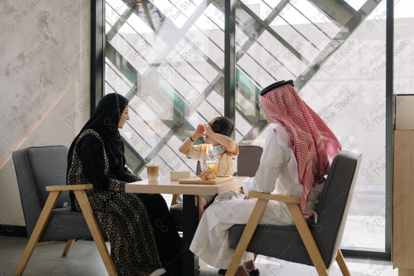 A family sitting at a table in a café; the mother is wearing a black abaya, the father is wearing a white thobe and ghutra, and the child is sitting between them.