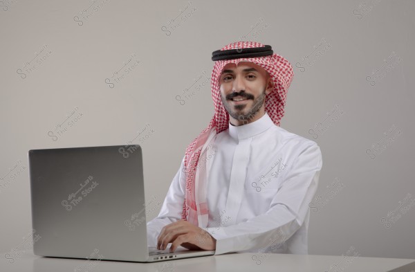 صورة لرجل جالس أمام كمبيوتر محمول. يرتدي الزي السعودي التقليدي, ثوب أبيض وشماغ أحمر وأبيض مع عقال أسود. يجلس الرجل مبتسماً في بيئة مكتبية بسيطة ذات خلفية رمادية.