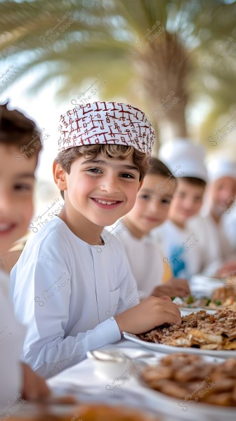 The image shows a group of children sitting around a table with various dishes of food. The main child in the image is wearing traditional Omani white clothing and an Omani traditional cap. There are palm trees in the background, indicating a sunny outdoor setting.
