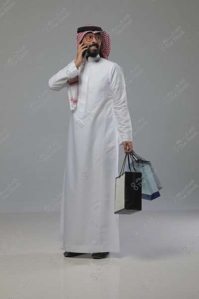 Image of a man wearing traditional Saudi attire, a white thobe with a red shemagh and a black agal. He is smiling as he holds a phone in his right hand and carries shopping bags of various colors in his left hand.