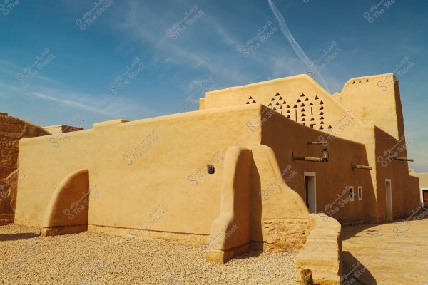 The image shows a traditional adobe building painted in a desert yellow color, featuring towers with triangular patterns and surrounded by gravel. The sky is blue and clear, adding a serene atmosphere to the scene.