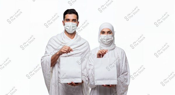 Image of a man and woman standing side by side, wearing white Ihram clothing. Both the man and woman are wearing white face masks and holding white paper bags. The background is white.