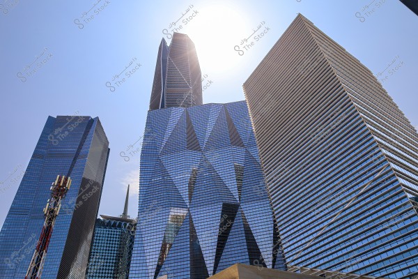 Image of a group of modern skyscrapers under a clear sky in daylight. The buildings feature reflective glass facades and various geometric designs, with sunlight visible behind them, enhancing the appeal of the scene. Communication antennas are visible atop one of the buildings.