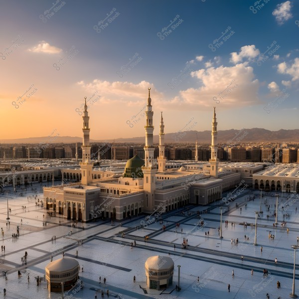 View of the Prophet\'s Mosque in Medina, Saudi Arabia. The image shows the large mosque with its distinctive green dome and four tall white minarets. The outer courtyard is spacious with people walking around. The background illustrates the city skyline and surrounding buildings under a blue sky with some white clouds, while the horizon is tinged with orange due to the sunset.