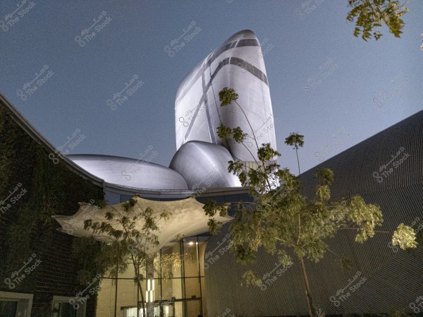 The image shows a modern building with an innovative and distinctive architectural design, illuminated at night, enhancing its beauty. The building is covered with shiny metallic materials and is surrounded by some green trees, with a prominent architectural canopy above a large glass entrance. The sky is clear and calm in the background.