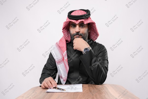 A portrait of a man sitting at a desk wearing a traditional black thobe with a red and white keffiyeh and a black agal. He is looking directly at the camera with one hand on his chin and the other arm on the table, holding a pen next to some papers. He is wearing a silver watch.