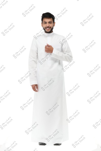 Image of a smiling man wearing a traditional white thobe, commonly associated with the Gulf region. The man is standing facing the camera against a clean white background. He is holding the thobe near his chest with his right hand.