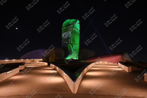 Nighttime image of the King Abdulaziz Center for World Culture (Ithra) in Saudi Arabia, illuminated in green with laser lights, displaying the Saudi flag. A straight pathway leading to the building is softly lit. A portion of the moon is visible in the dark sky.