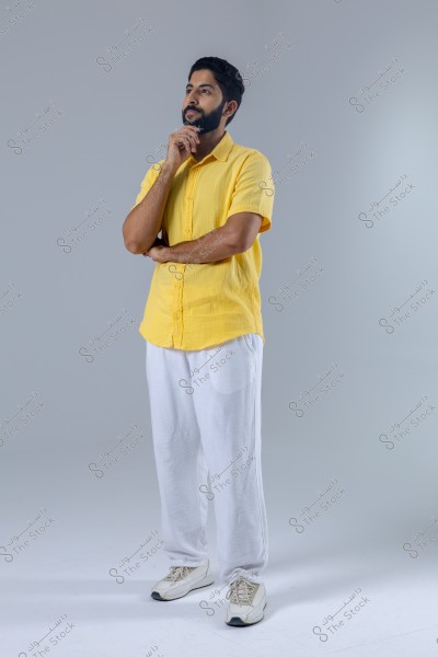 A portrait of a man standing in a thinking pose with his hand on his chin. He is wearing a short-sleeve yellow shirt, white pants, and white sneakers. The background is gray.