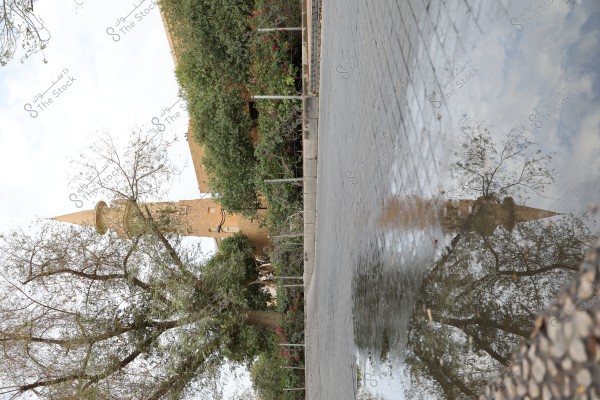 An image of a traditional mud brick tower surrounded by green trees and walls, with its reflection visible in a puddle on a tiled ground. The weather appears cloudy with a slight view of the sky. The scene evokes a sense of heritage and traditional architecture.
