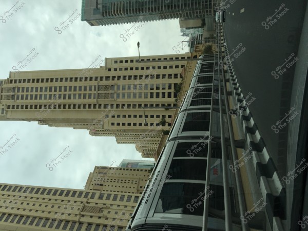 Skyscrapers under a cloudy sky in an urban scene. The buildings are tall and modern with glass facades, and there is a pedestrian bridge visible in the foreground. The road is wide and runs close to the buildings.