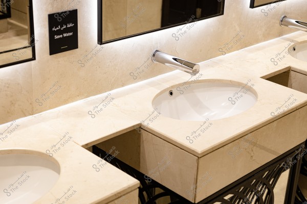 صورة لحوض غسيل حديث بلون بيج, مع صنبور معدني مصقول. توجد مرايا كبيرة خلف الحوض وإضاءة مخفية تضيف لمسة أنيقة. يوجد لافتة باللغة العربية والإنجليزية مكتوب عليها "حافظ على المياه" مثبتة على الجدار.
