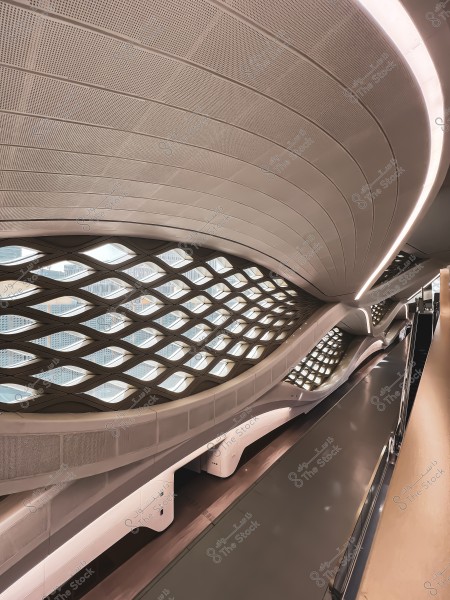 Image of a modern train station with a unique architectural design. The wavy ceiling features integrated lights and triangular windows that allow natural light in. The floor is clean and shiny with an escalator on the right side. The design appears futuristic with a sleek and streamlined aesthetic.