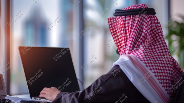 An image of a man wearing traditional Saudi attire, including a thobe, ghutra, and agal, sitting at a laptop in a modern office setting with a window overlooking an urban view in the background.