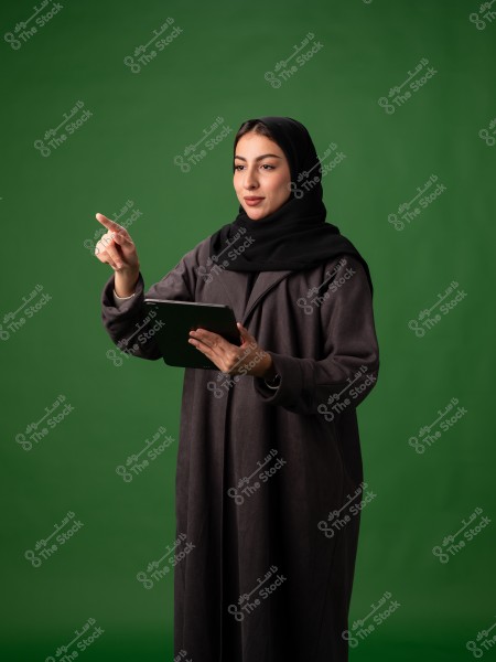 امرأة ترتدي عباءة سوداء وحجابًا تقف أمام خلفية خضراء. تبدو أنها تتفاعل مع جهاز لوحي بيدها اليسرى وتشير بإصبعها الأيمن. ترتدي ملابس تقليدية تشير إلى بلد شرق أوسطي.
