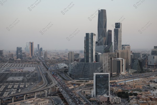 The image shows an aerial view of the skyline of Riyadh, Saudi Arabia. It features several modern and architecturally diverse towers, prominently including the Kingdom Tower and Al Faisaliah Tower. Highways intersect among the buildings, with visible traffic on the city streets. The background shows various shades of grey in the sky.