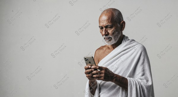 An elderly man with a white beard wearing a white cloth similar to an izar, looking at a smartphone in his hands. He stands before a plain light gray background. His expression is serious and focused, with lighting highlighting his facial features and attire.