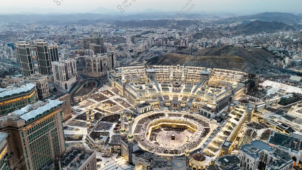 Aerial view of the Great Mosque of Mecca in Saudi Arabia. The Kaaba is visible in the center of the main courtyard, surrounded by large numbers of worshipers. Modern buildings and tall hotels surround the mosque, with mountains visible in the distant horizon.