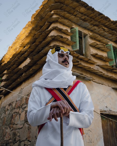 A snapshot of a Saudi man wearing traditional costume on the founding day, standing in front of a mud building and leaning on a stick, traditional costume, foundation day, ancient ancient buildings