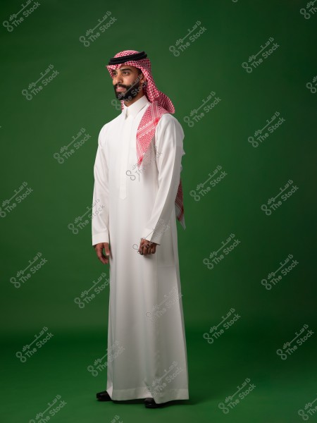 A portrait of a man wearing traditional Saudi attire, consisting of a white thobe, a red checkered shemagh, and a black agal. The man is standing in a side pose against a green background.