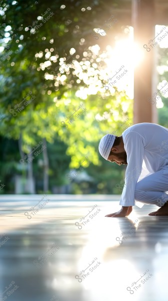 Image of a man wearing a white robe and a white prayer cap, positioned in a bowing or prayer stance in an open space. The lighting in the background provides a serene atmosphere with sunlight streaming through green trees, adding a touch of tranquility and calmness to the scene.