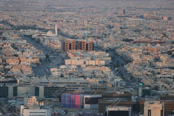 An image of a large city sprawling across a vast area, showcasing tightly packed buildings arranged along organized streets. A prominent mosque with a large white dome and minaret stands out amidst the other structures. The cityscape suggests a modern environment in development. Soft lighting adds a lively touch to the streets winding through the buildings.