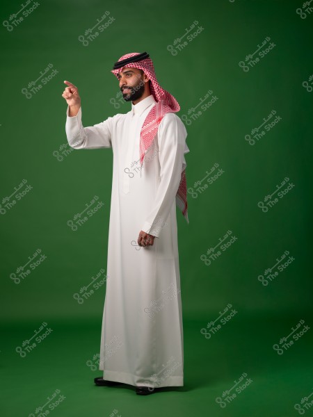 An image of a man wearing a white thobe, a red-checkered ghutra, and a black agal standing against a green background. He appears to be pointing with his hand to the right. The attire reflects traditional Gulf culture.