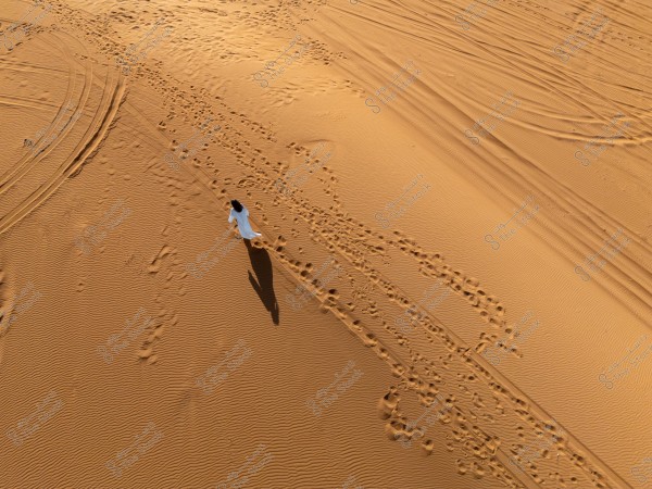 Aerial view of a person wearing a white garment walking in the desert, leaving a clear trail on the orange rippled sand. Wheel tracks are also visible on the sand.