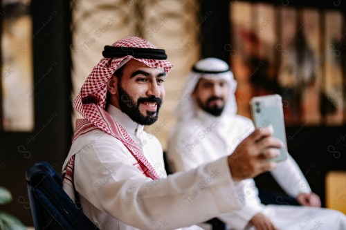 شاب سعودي يرتدي الزي السعودي التقليدي يرفع هاتفه لالتقاط صورة شخصية مع صديقه في المقهى