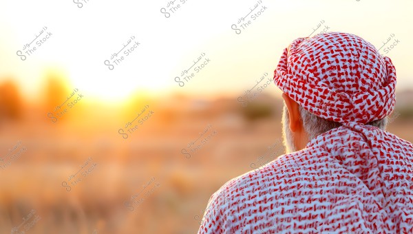 Image of a man wearing a traditional white and red checkered headscarf, looking towards the sunset in a desert-like natural background. The photo shows the back of the man with emphasis on the fabric\'s details and traditional style.