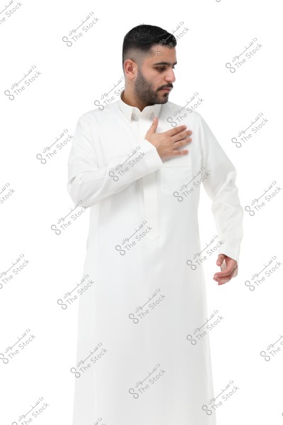 A portrait of a man wearing a long white thobe, looking to the side, with his left hand placed on his chest. The background is completely white, highlighting the details of the traditional clothing. The thobe suggests traditional attire from the Arabian Gulf region, such as Saudi Arabia.