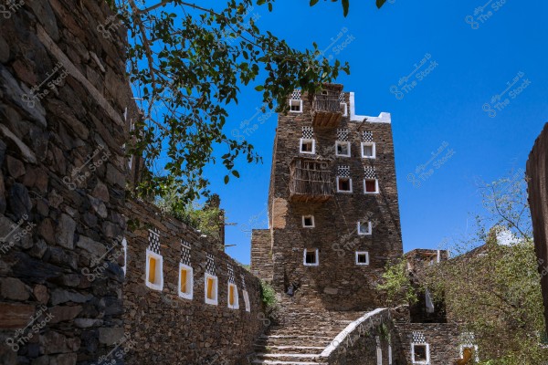 The image shows a traditional building made of stone and clay with small square windows framed in white, featuring wooden balconies. The sky is clear blue, and some green branches appear at the top of the image. There\'s a stone wall extending on the left side with windows decorated with white frames and a yellow center.