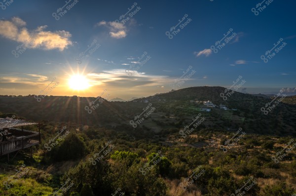 منظر طبيعي يظهر غروب الشمس خلف الجبال مع سماء زرقاء صافية وسحب قليلة ومنزل في الأفق.