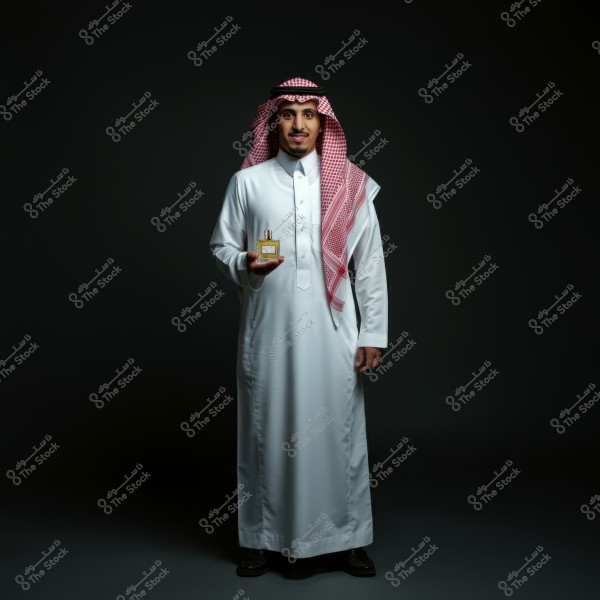 An image of a man standing in a dark setting, wearing traditional Saudi attire known as a white thobe, red checkered ghutra, and black agal. The man is holding a perfume bottle in his right hand and appears to be smiling.