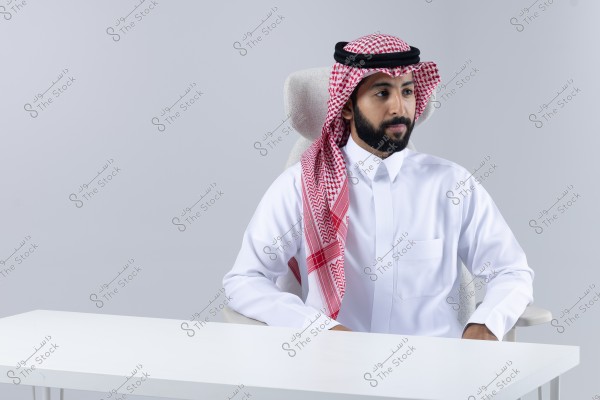 Image of a man sitting on a chair behind a white table. He is wearing a white thobe and a red and white shemagh with an agal, which is traditional attire commonly seen in Saudi Arabia and the Gulf countries. The background is simple and monochromatic.