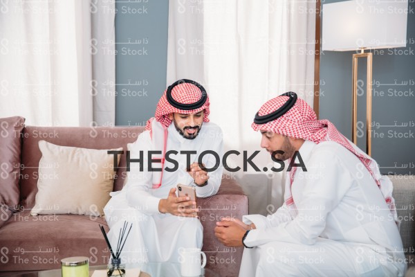 رجلان سعوديان يرتديان الثوب و الشماغ، يجلسان في المجلس و ينظران الى الجوال