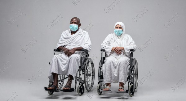 The image shows two individuals seated in wheelchairs, both wearing white Ihram clothing and face masks. The person on the left wears the Ihram without a head covering, while the person on the right is wearing a headscarf. They appear to be in a neutral, reflective environment.