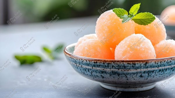 An image of round orange sorbet balls arranged in a ceramic bowl with blue and white detailing. The bowl sits on a smooth gray surface, and the sorbet balls are garnished with fresh green mint leaves, adding a touch of elegance and nature. A blurred background with additional green leaves is visible.
